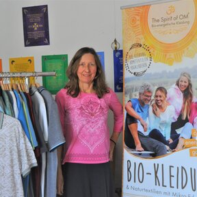 Inhaberin des Unternehmens "Ökologische Wellnessbekleidung" in ihrem Laden umgeben von Kleidung und eines Werbeplakats.