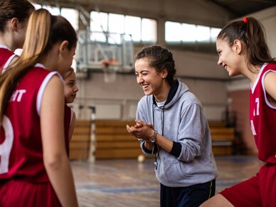 gemeinsamhelfen Vier Mädchen in Sport-Trikots stehen in einem Halbkreis um ihre Trainerin herum.