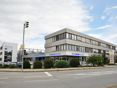 Nussbaum Medien Weil der Stadt Kastenförmiges, graues Gebäude hinter einer Straße, am Gebäude eine Banderole mit dem Schriftzug "Nussbaum Medien".