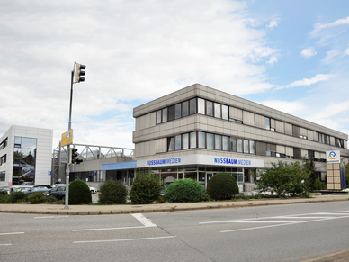 Kastenförmiges, graues Gebäude hinter einer Straße, am Gebäude eine Banderole mit dem Schriftzug "Nussbaum Medien".