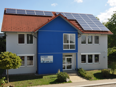 Nussbaum Medien Dußlingen Ein hellblaues Haus mit braun-rotem Dach und Solaranlage auf dem Dach. In der Mitte ein dunkelblauer Erker, im unteren Teil ein Schild mit dem Logo und Schriftzug von Nussbaum Medien.