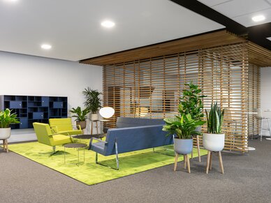 Ettlingen, Standort, Bürogebäude, Nussbaum medien Sitz-Lounge mir grünen und grauen Lounge-Möbelstücken, einem grünen Teppich, Holzverkleidung an der Wand und verschiedenen Pflanzen auf Hochständern aufgestellt.